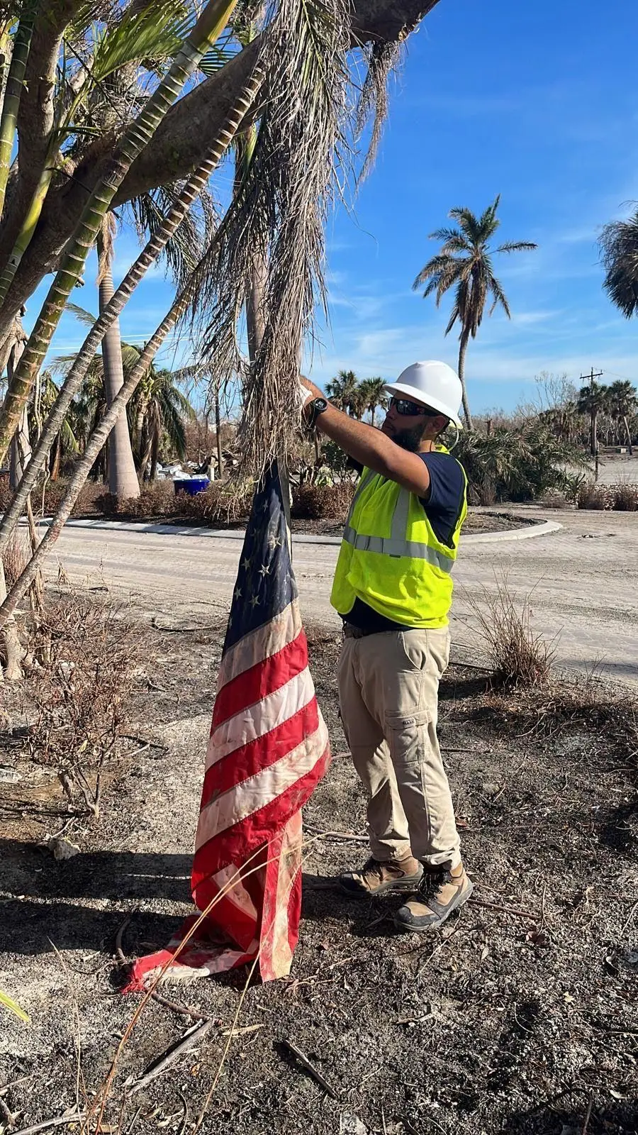 A crew member in a hard hat and safety vest freeing a damaged American flag from a palm tree after a storm