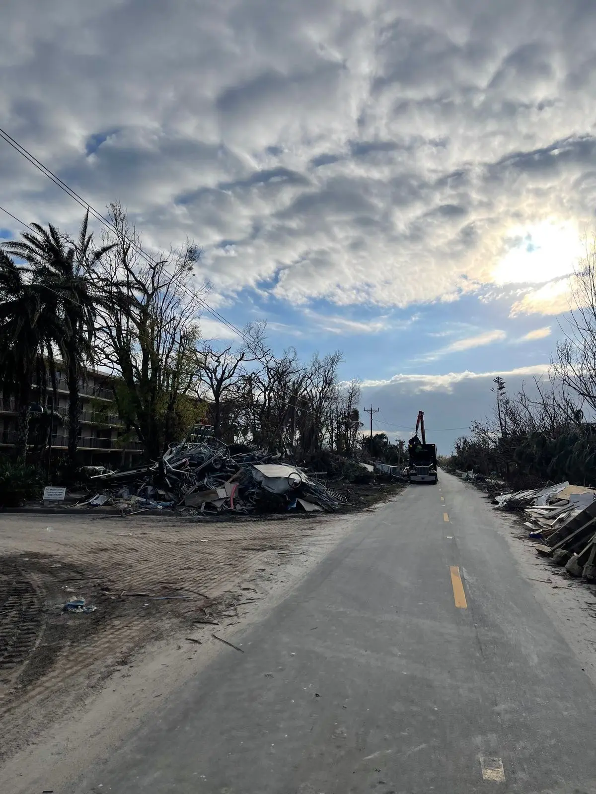 A street lined with storm debris and a grapple truck loading it under a cloudy sky