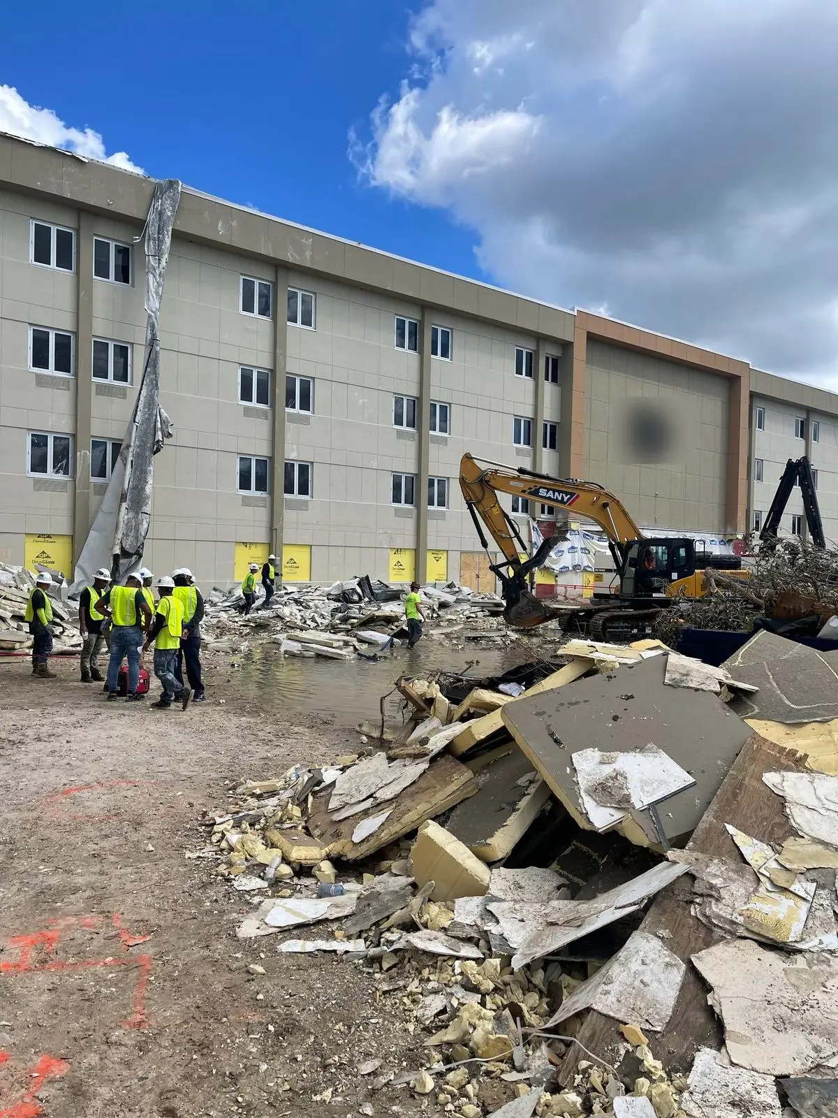 Crews in hard hats and safety vests beside an excavator and a pile of debris at a hospital after hurricane damage