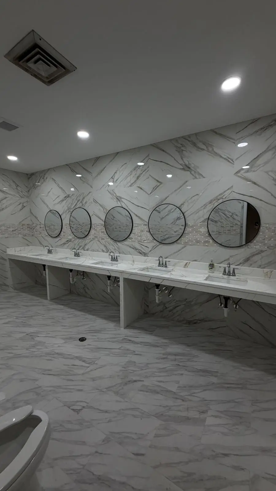 Renovated restroom with marble-look tile walls and floor, round black mirrors and a long vanity