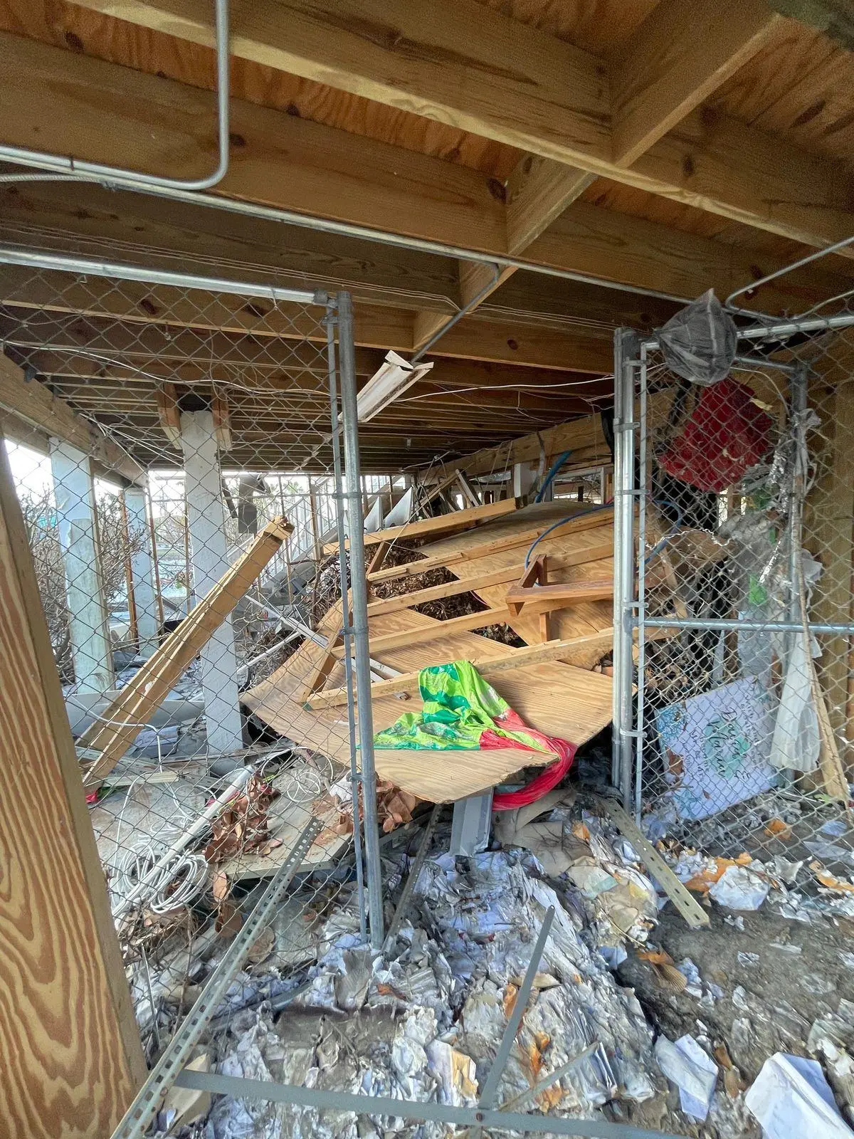 Storm debris and broken lumber beneath a raised house, behind chain-link fencing